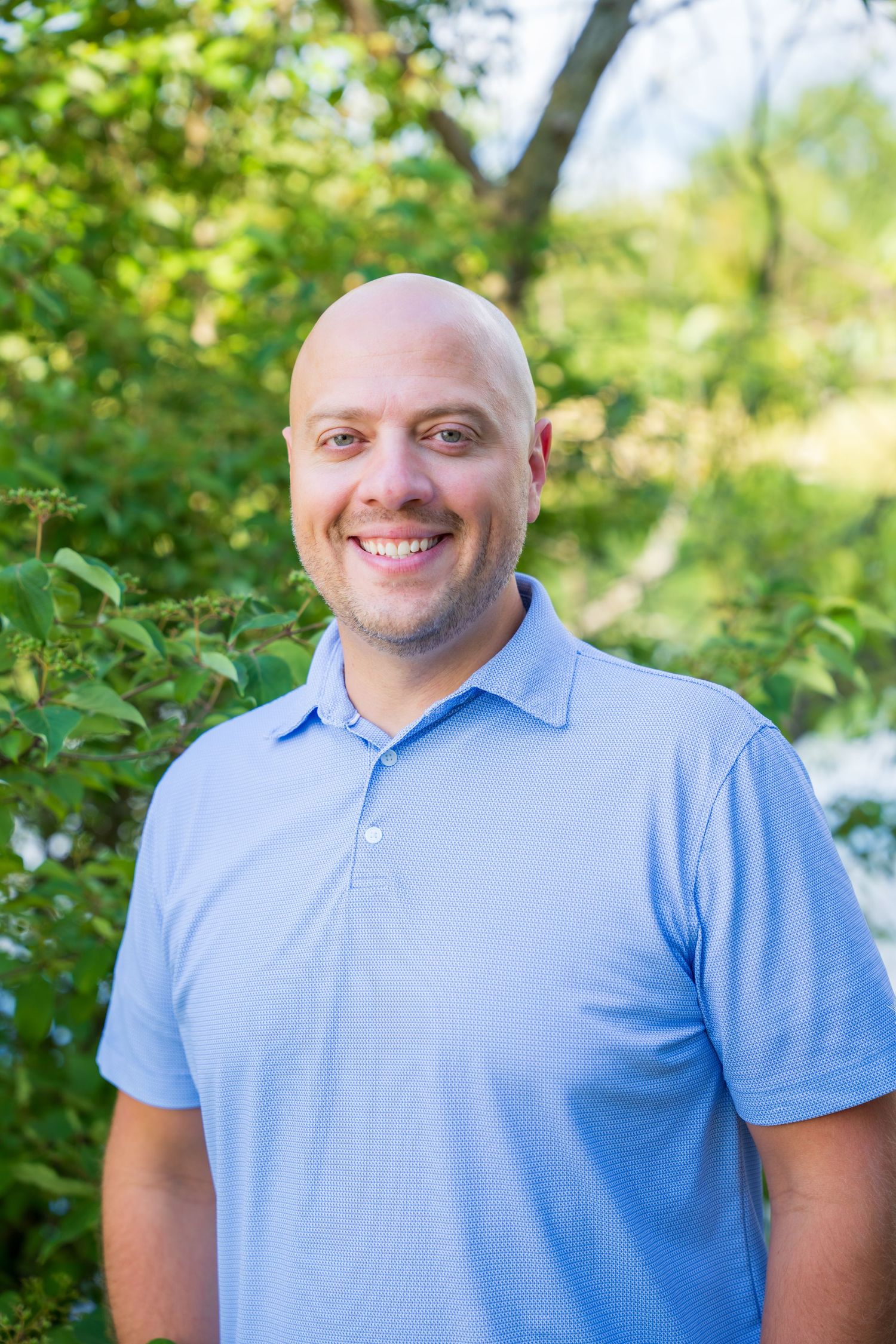 Smiling bald man in a light blue polo shirt posing outdoors with green trees and foliage in the background.