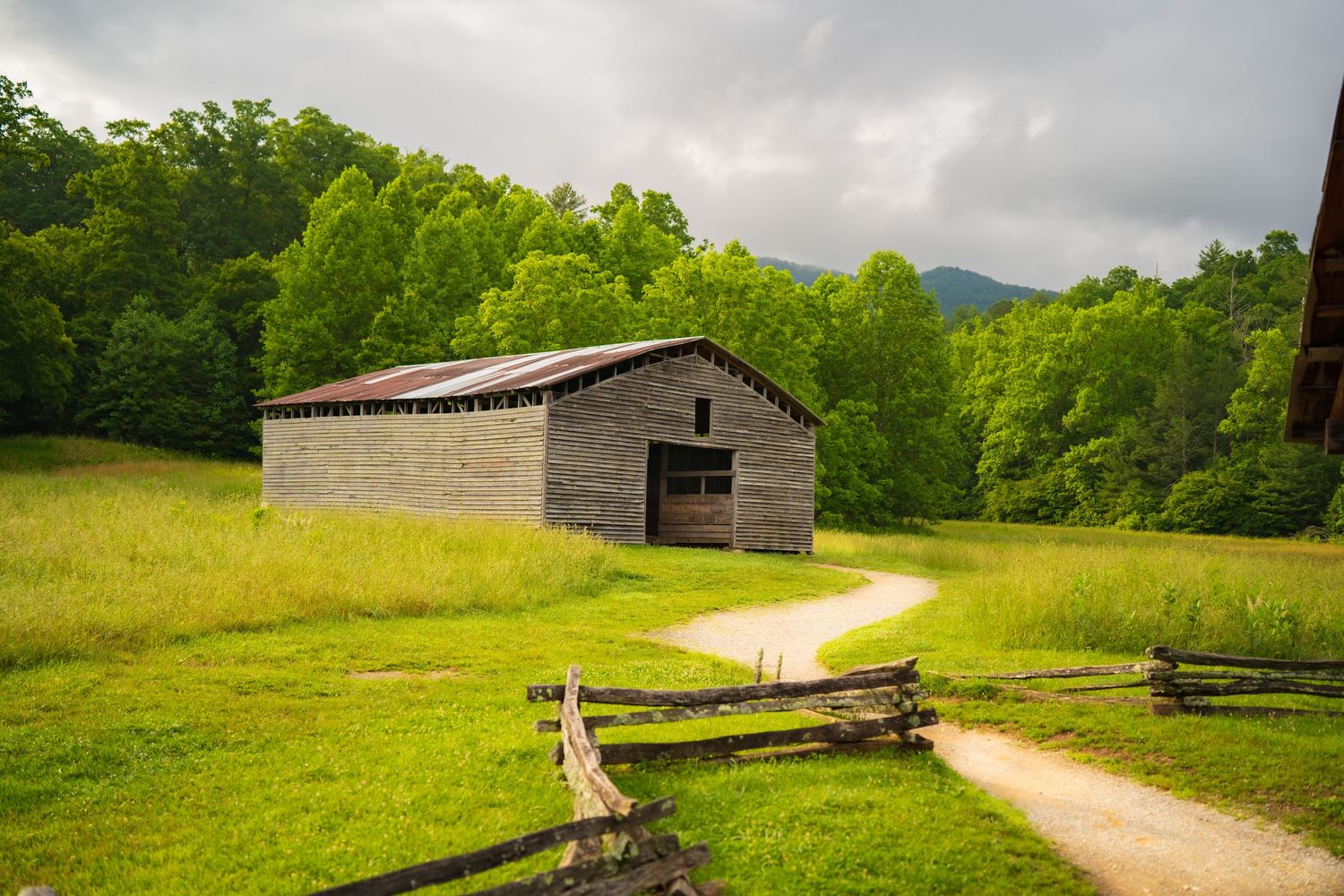 Rustic wooden barn beside a winding dirt path in a lush green valley with forested mountains in the background.