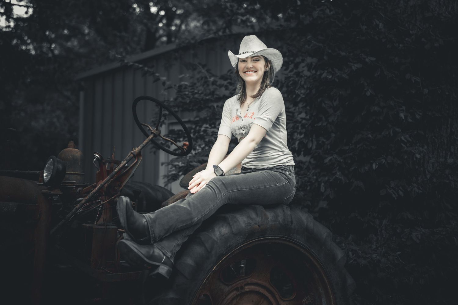 Smiling woman in white cowboy hat sitting on vintage rusty tractor wearing jeans and boots in rustic outdoor setting.