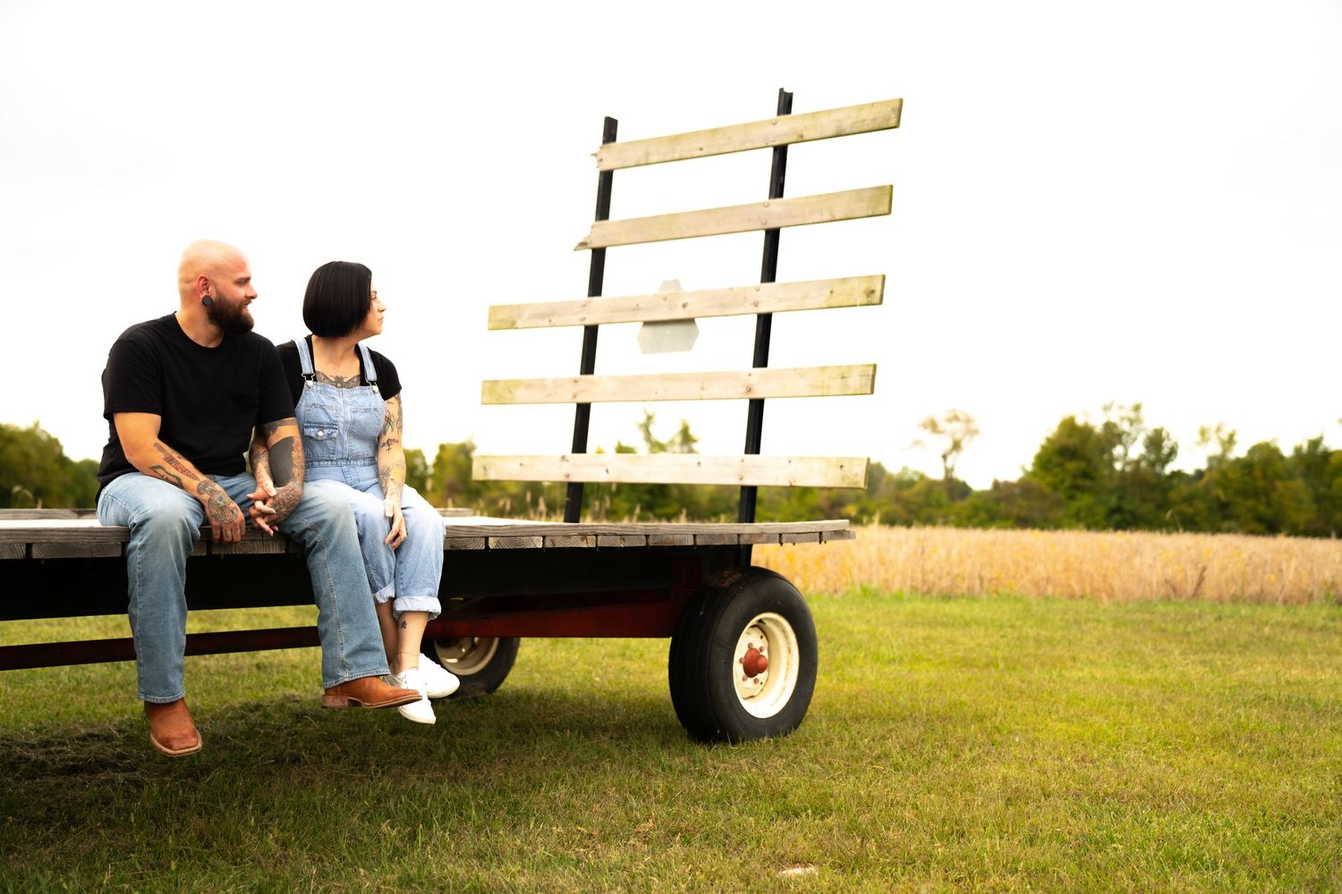 Couple sitting together on a wooden farm wagon in a green field, sharing a tender moment outdoors.