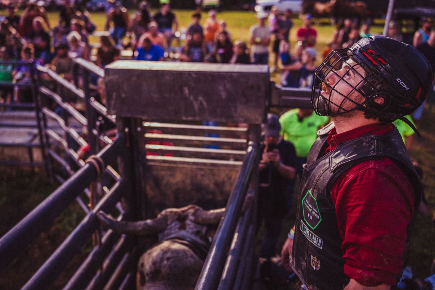 A rodeo rider in helmet and vest prepares near metal gates as a crowd watches at an outdoor bull riding event.