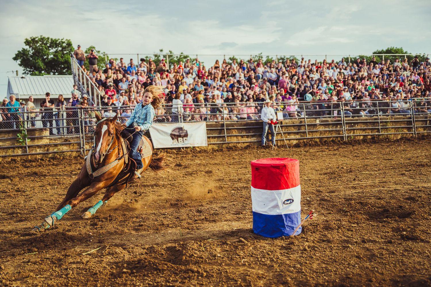 A barrel racer on a brown horse rounds a red, white, and blue barrel at a packed rodeo arena.