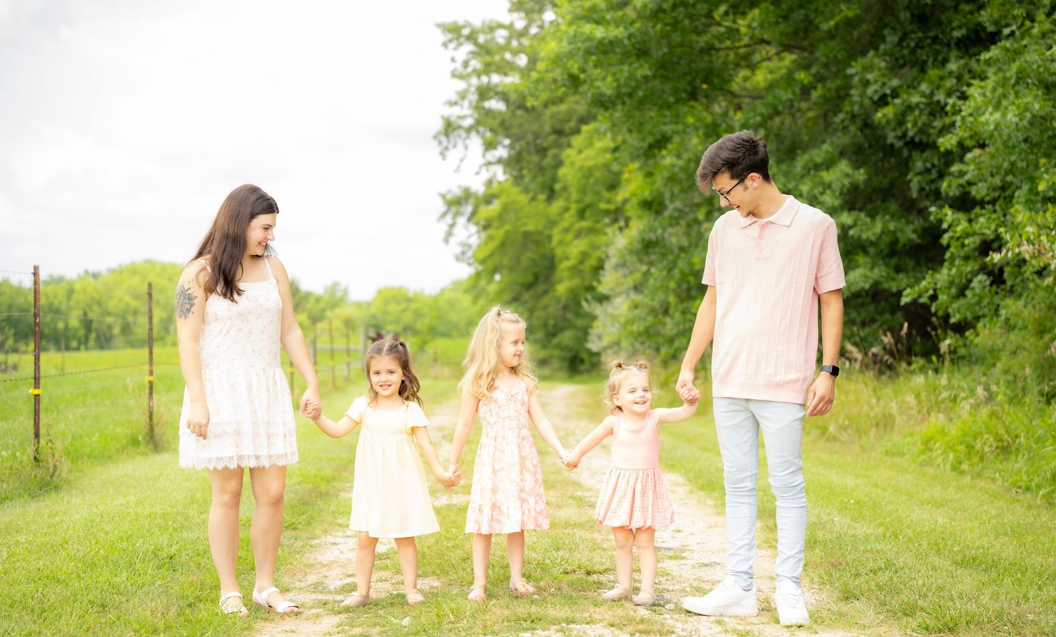 Family of five holding hands on a grassy path surrounded by lush green trees on a sunny day.
