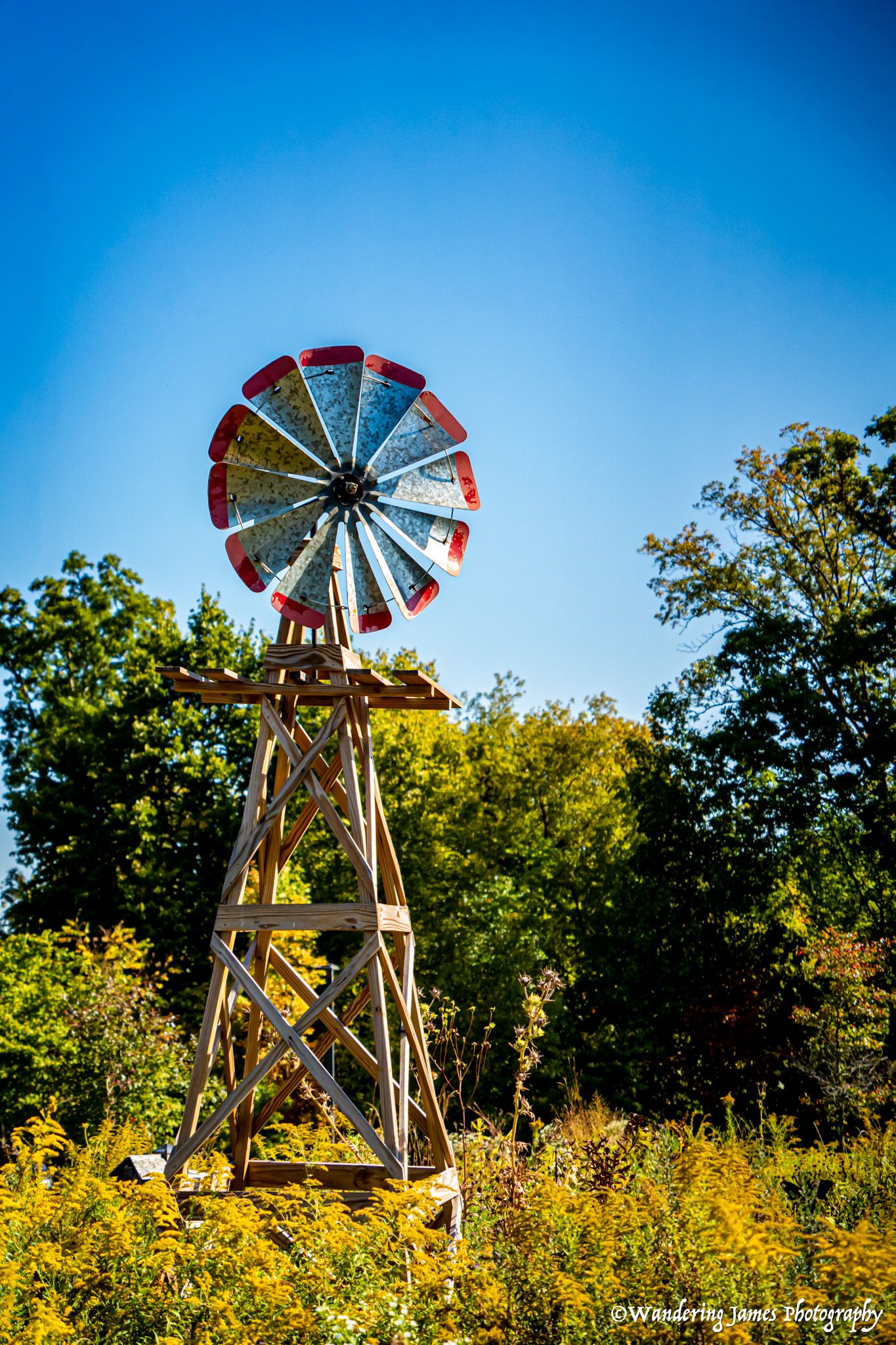 Colorful vintage wooden windmill with red-tipped blades against a clear blue sky surrounded by trees.