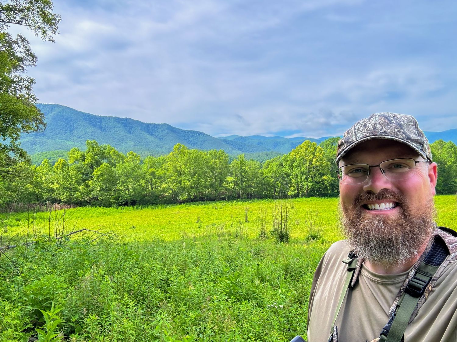 Smiling bearded man in camo cap taking selfie with lush green mountain meadow and misty blue mountains in background.