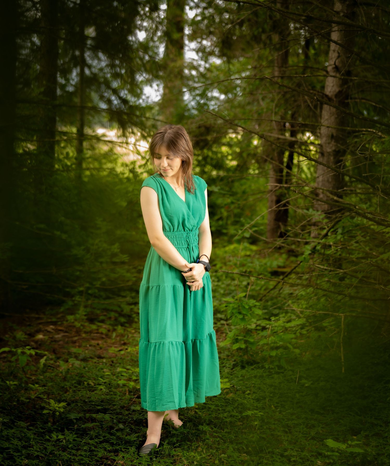 A woman in a green midi dress stands thoughtfully in a lush forest setting.