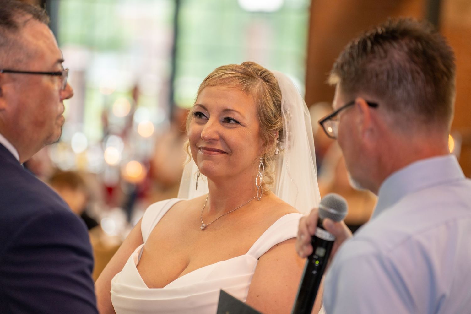 Smiling bride in white gown and veil listens during wedding ceremony indoors.