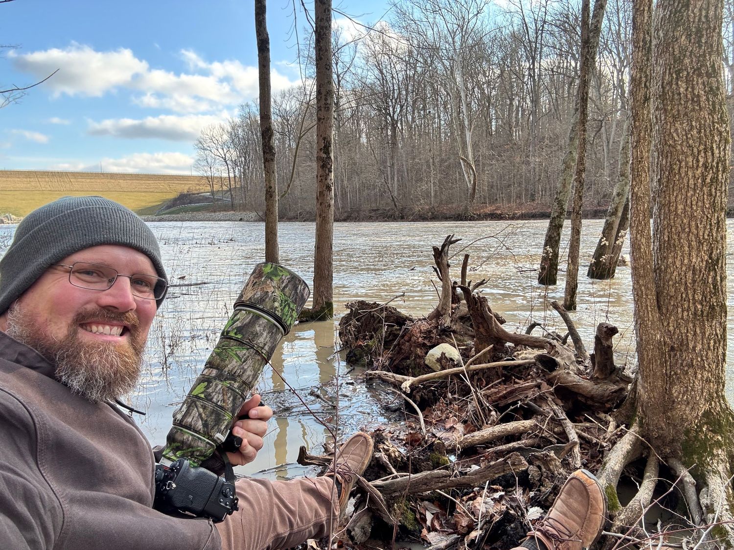 Bearded man in camo gear smiling near a flooded river with fallen tree roots and bare winter trees in background.