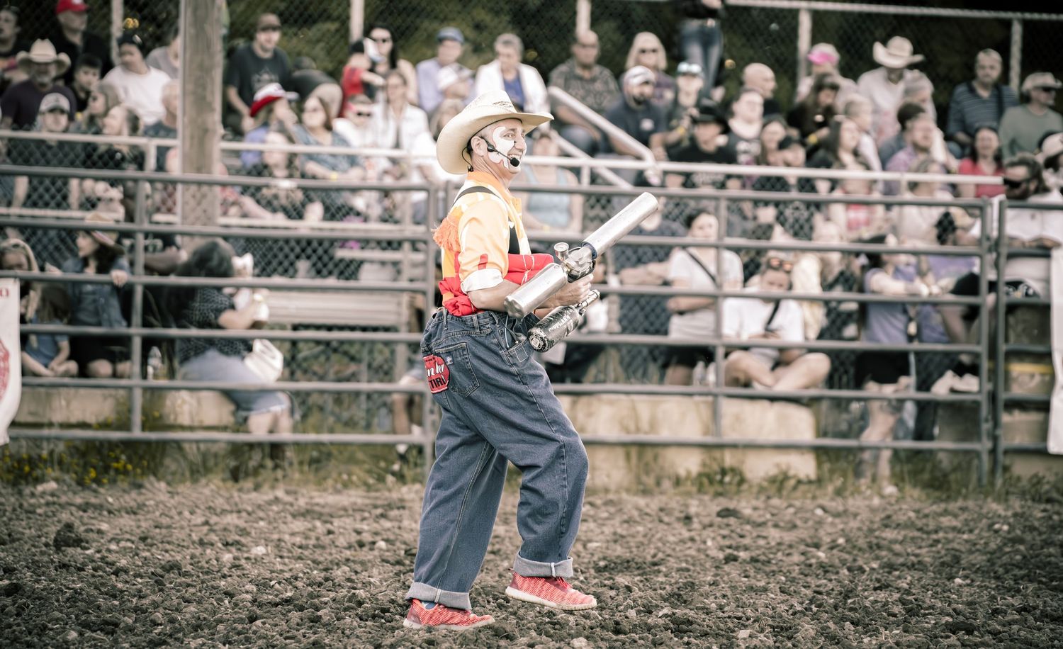 Rodeo clown in colorful costume and helmet entertains the crowd in a dirt arena.