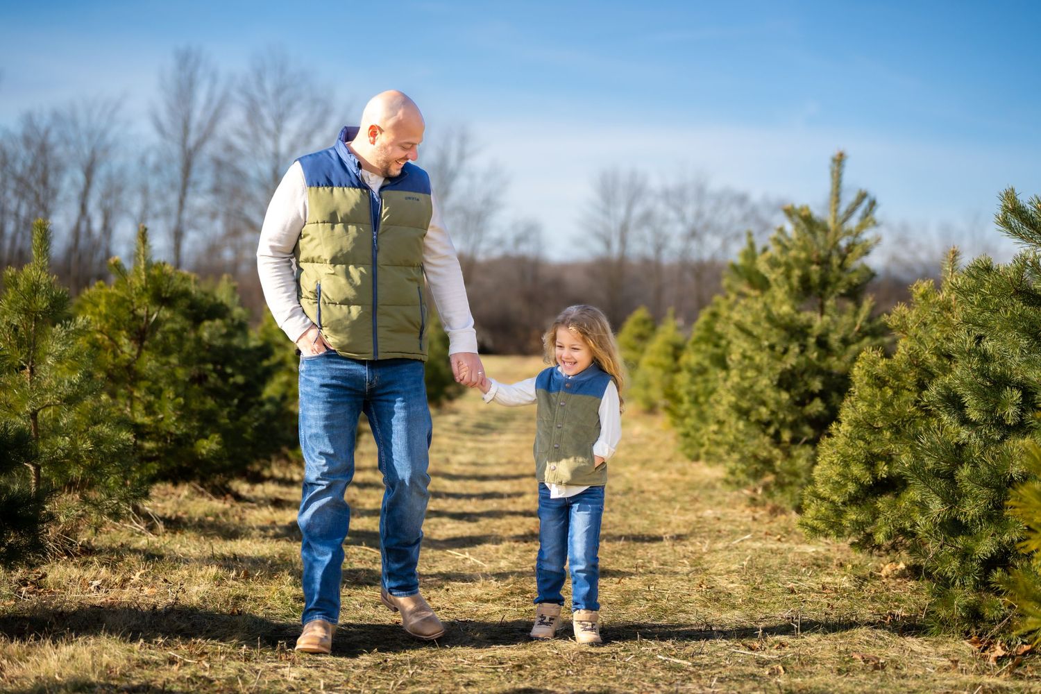 A grandfather and young girl walk hand-in-hand through rows of Christmas trees on a sunny farm.