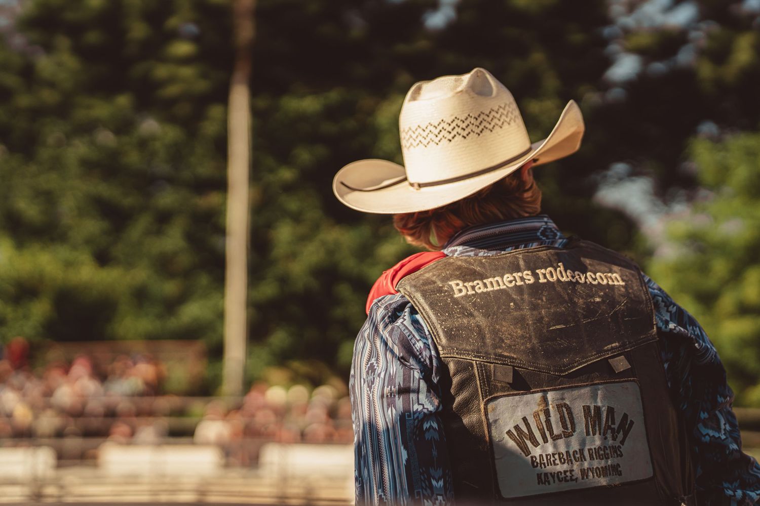 A cowboy wearing a straw hat and Wild Man Bramers rodeo jacket stands at a rodeo event.
