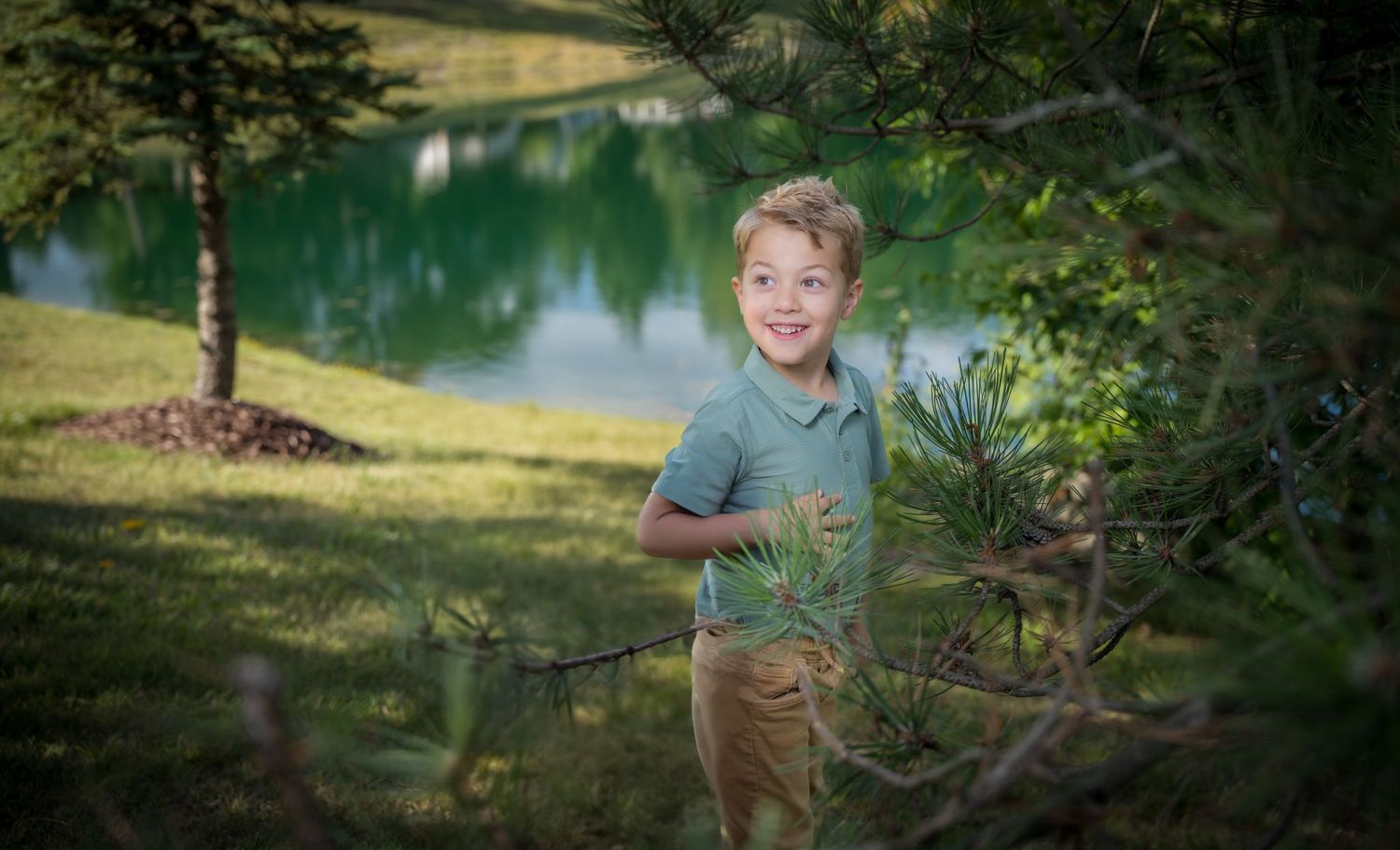 Smiling young boy standing among pine branches with a serene green lake and trees reflected in the background.