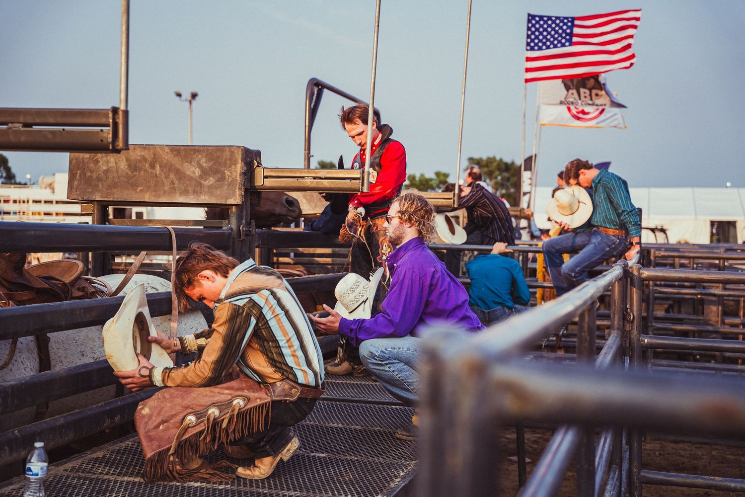Cowboys prepare behind the chutes at a rodeo event with an American flag waving in the background.