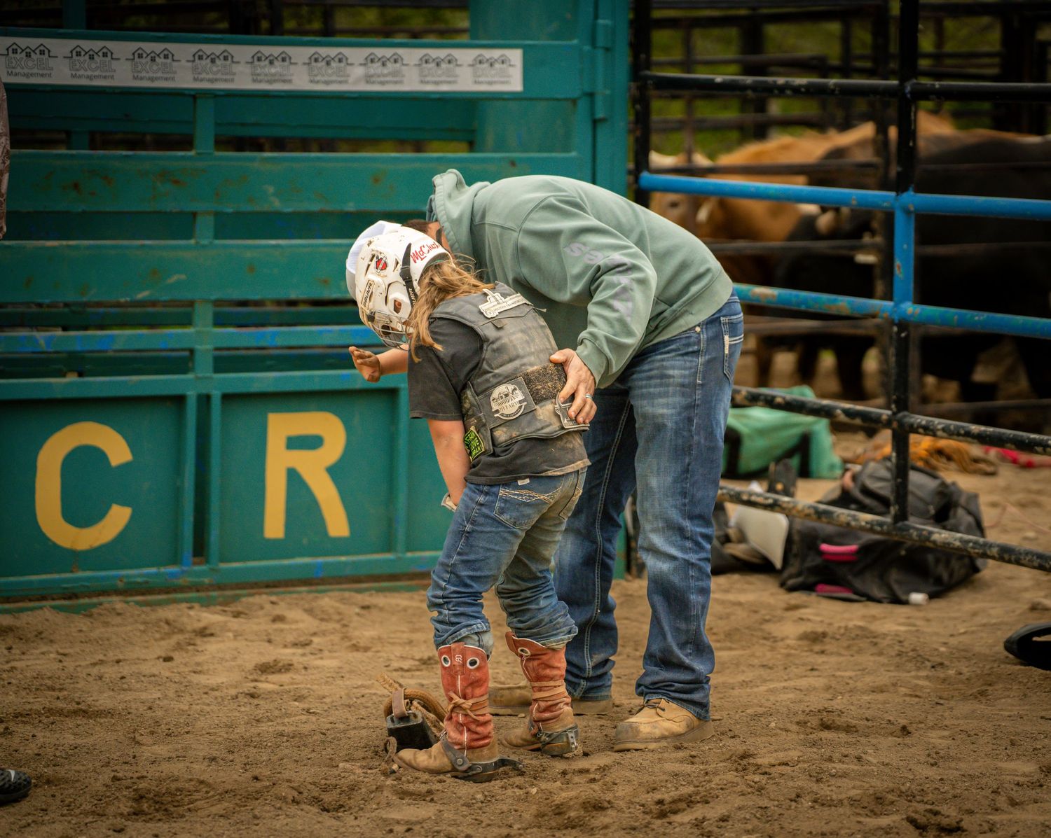 A rodeo helper assists a young rider with gear adjustment in the dirt arena near the chute.