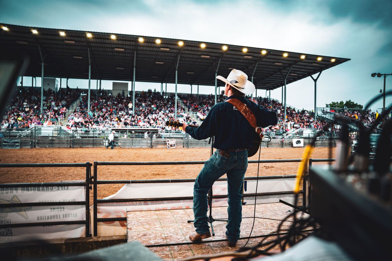 Cowboy in white hat stands at rodeo arena fence, watching competition with packed grandstands in background.