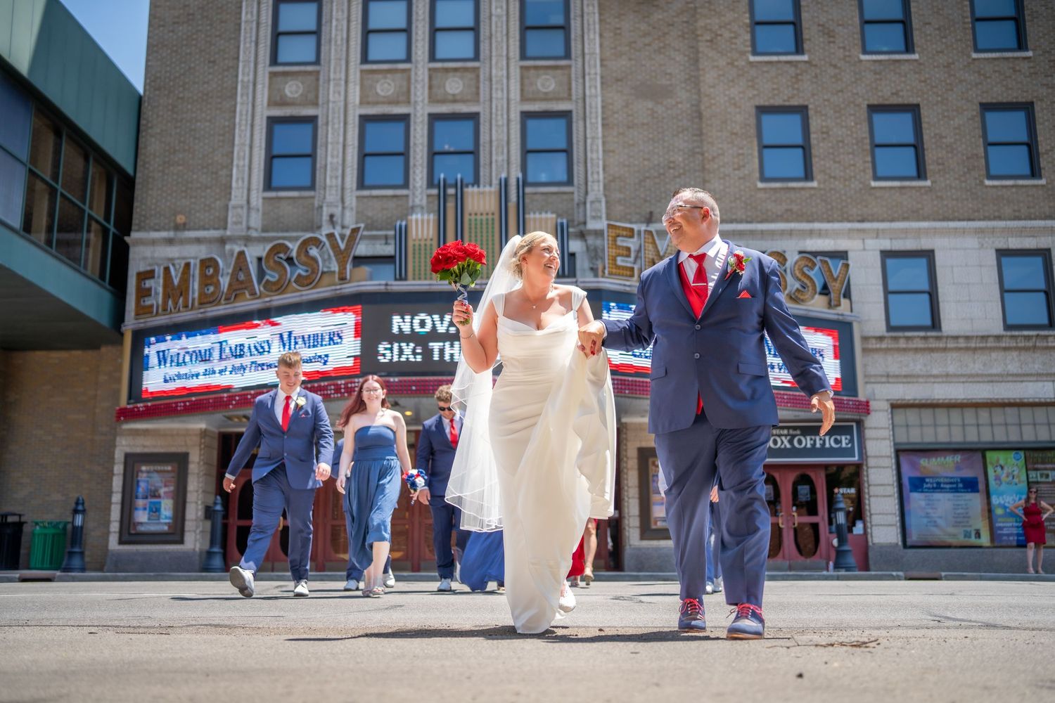 Wedding party walks outside the Embassy Theatre in formal attire, bride in white gown with groom in red tie.