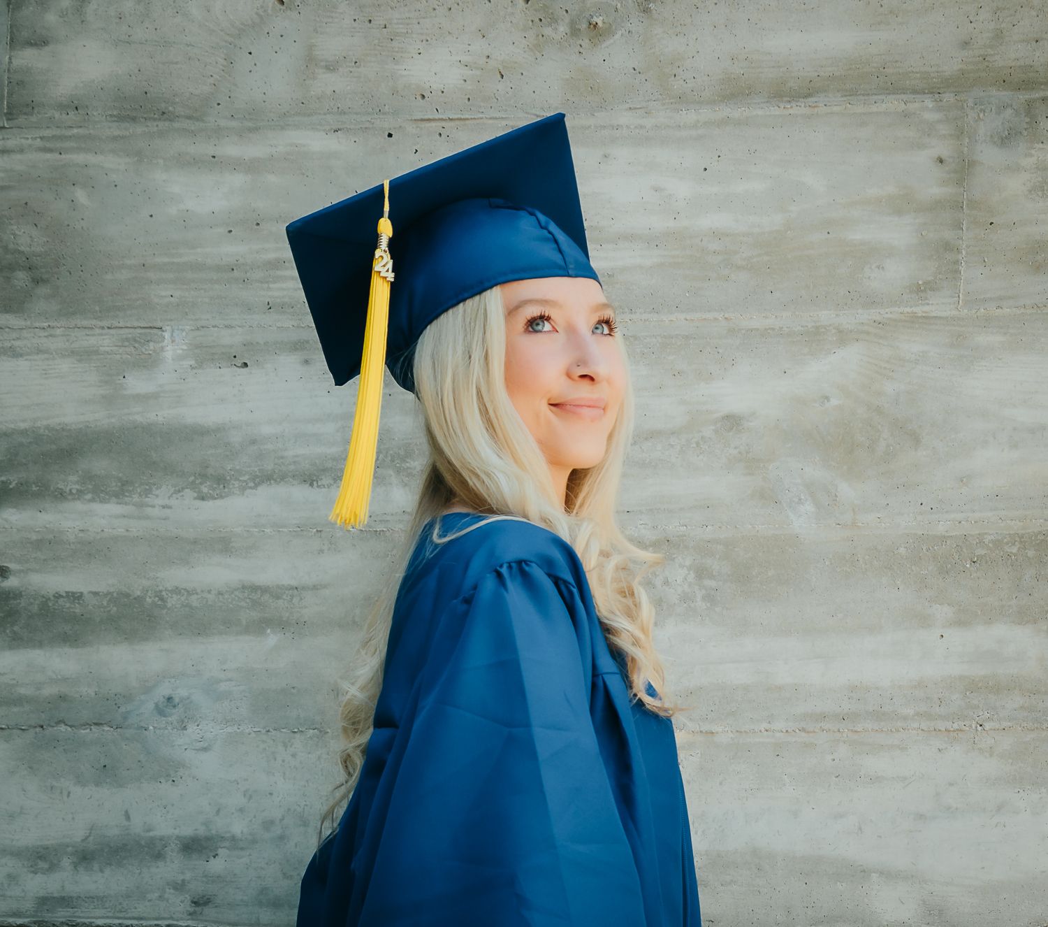 Blonde graduate in blue cap and gown posing in profile against concrete wall.