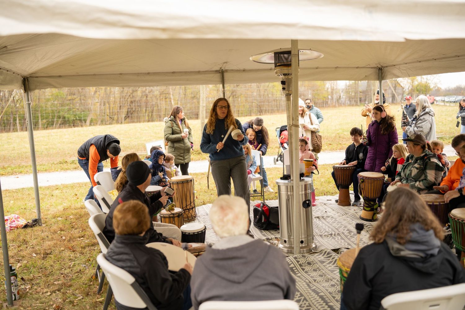 Group of people participate in a drum circle under a white tent canopy in an outdoor fall setting.