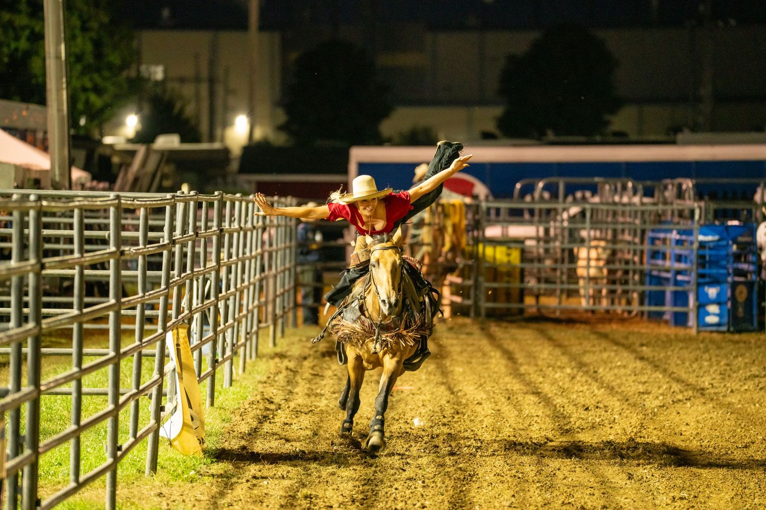 Rodeo rider on a bucking bronco at a nighttime rodeo event near metal fencing.