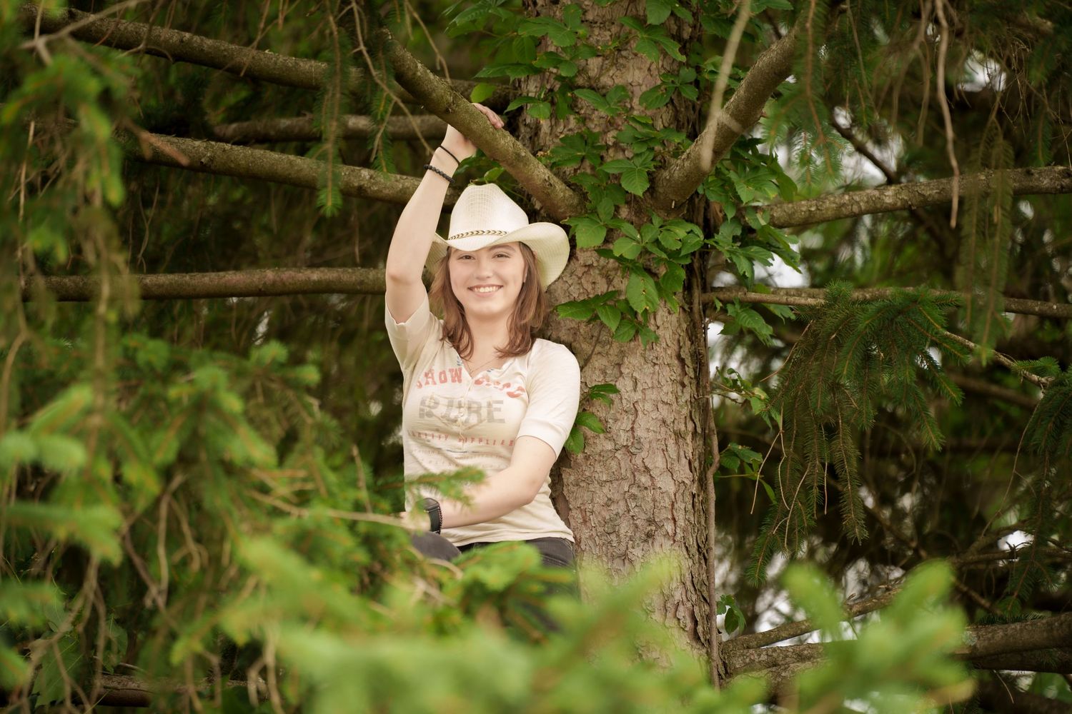 A smiling woman in a white cowboy hat poses among lush green tree branches outdoors.