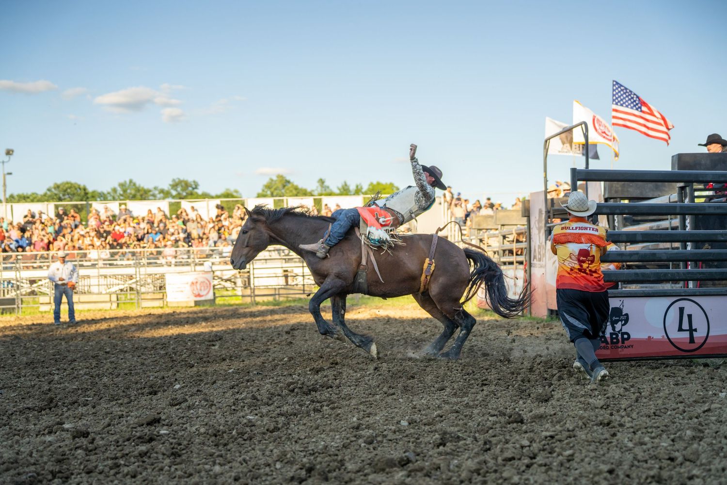 Rodeo cowboy riding a bucking brown horse near the gate at a sunny outdoor rodeo competition.