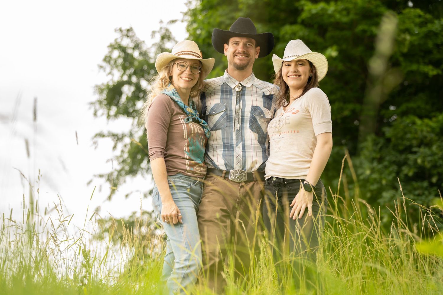 Three friends in cowboy hats smile outdoors near a lake surrounded by lush green grass and trees.