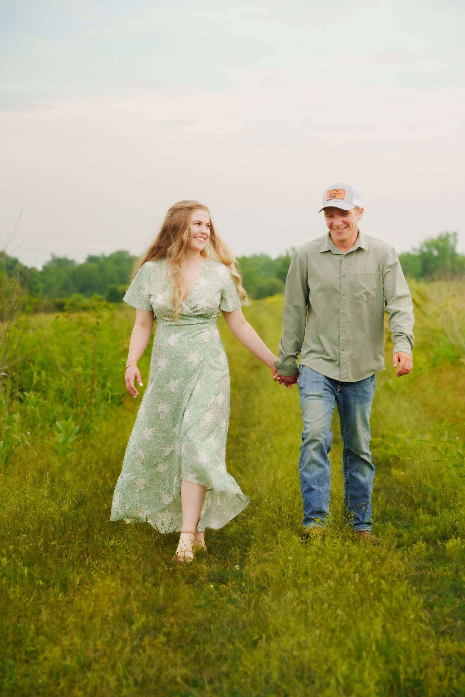 A smiling couple walks hand-in-hand through a lush green field, woman in floral dress, man in jeans and cap.