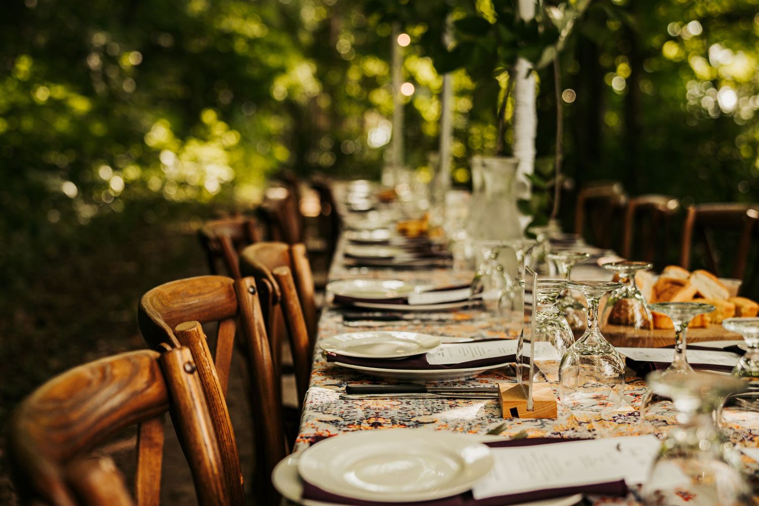 Elegantly set long outdoor dining table with wooden chairs, white plates, and glassware in a forest.