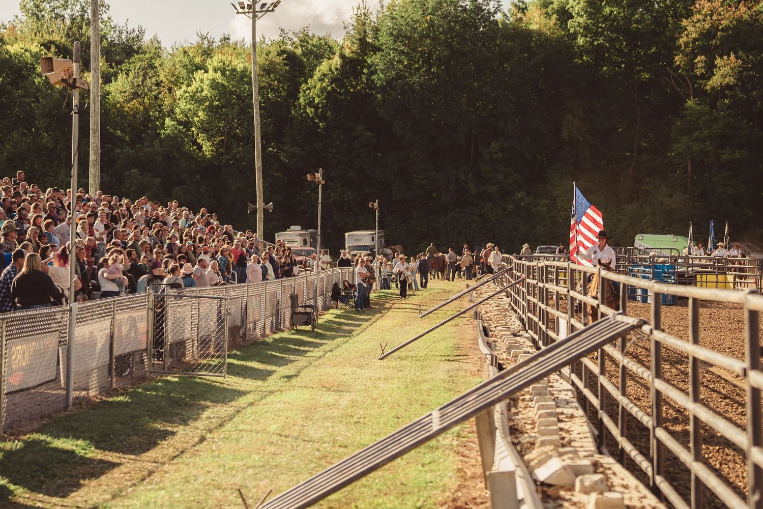 Large crowd of spectators lines the fence at an outdoor rodeo arena with an American flag on display.