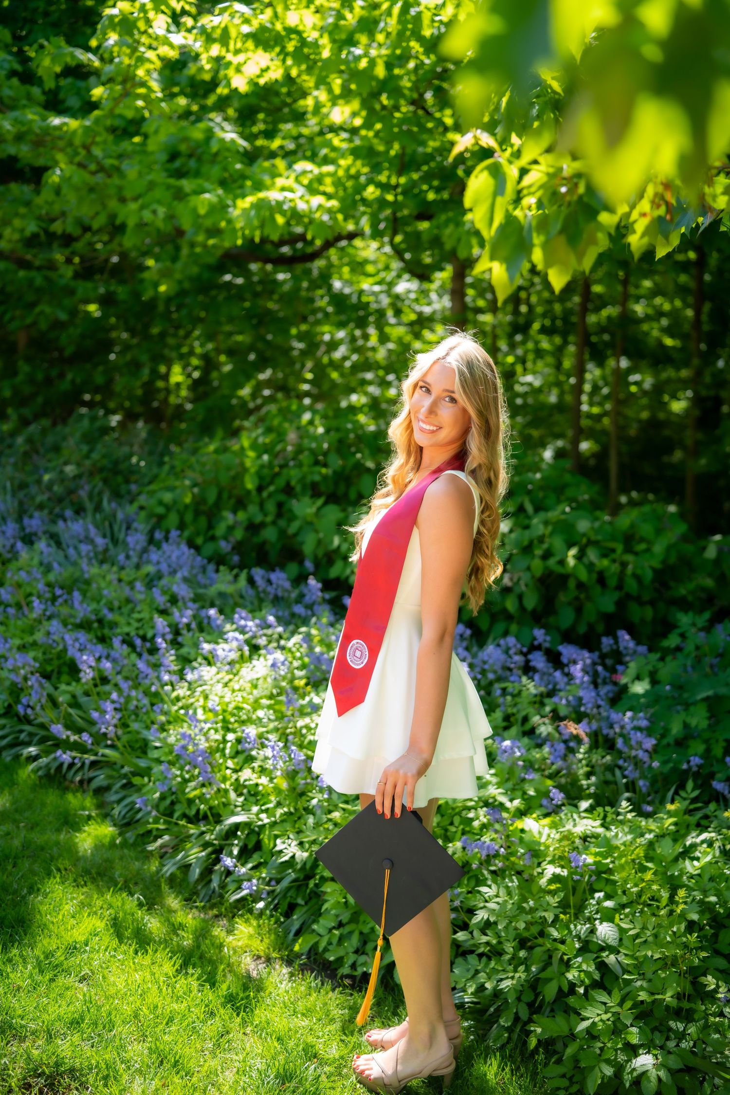 Blonde graduate in white dress holding cap with red stole standing in lush green garden.