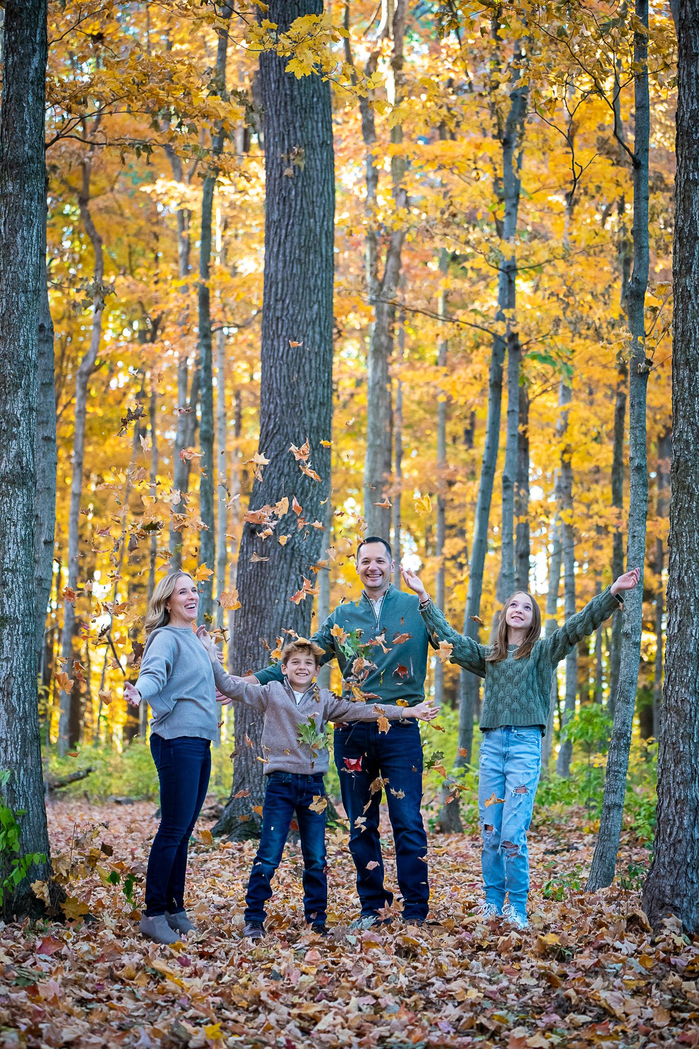 A cheerful family of four tosses autumn leaves in a golden fall forest with tall trees.