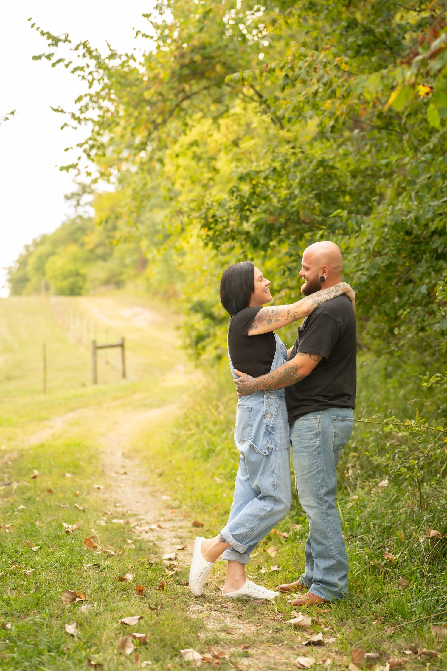 Couple embracing on a rural dirt path surrounded by green trees in soft autumn light.