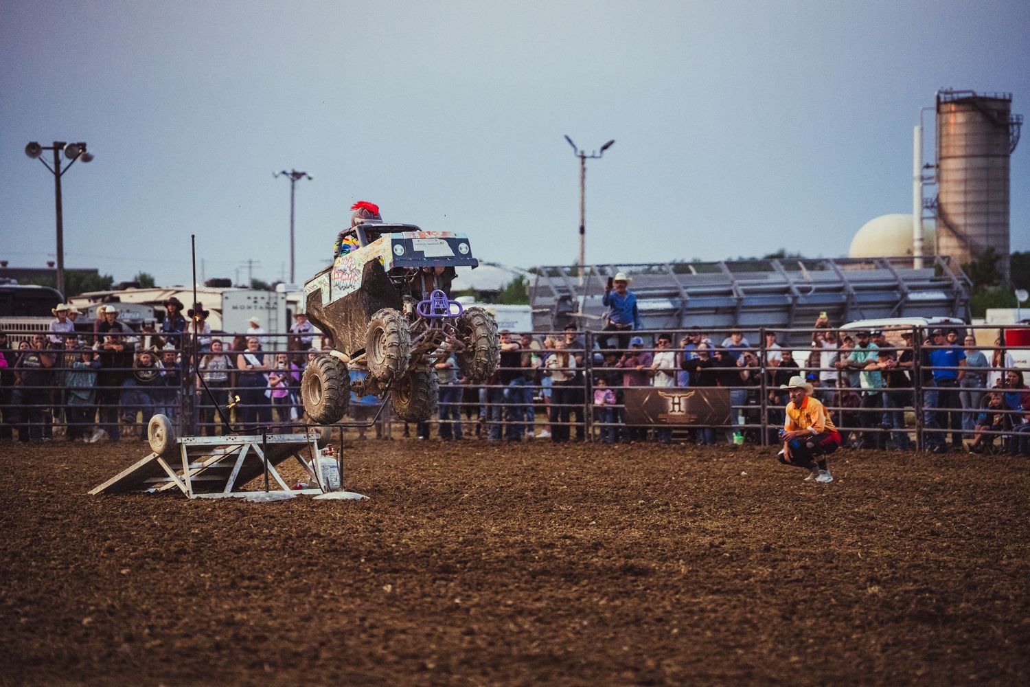 A monster truck soars over a jump at an outdoor rodeo arena packed with spectators under a clear sky.