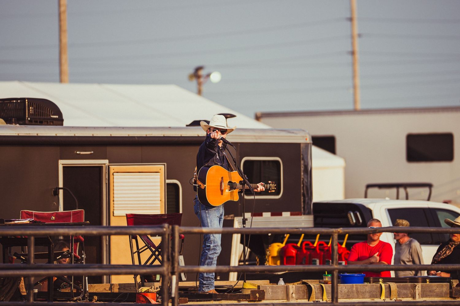 Country musician in cowboy hat plays acoustic guitar on outdoor stage at a rodeo event with trailers in background.