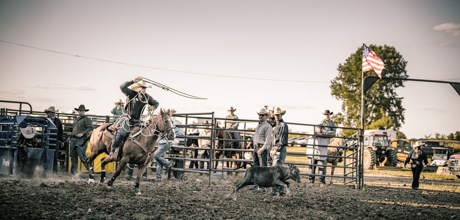 Cowboys compete in a rodeo roping event, with a rider on horseback chasing a calf in a dirt arena.