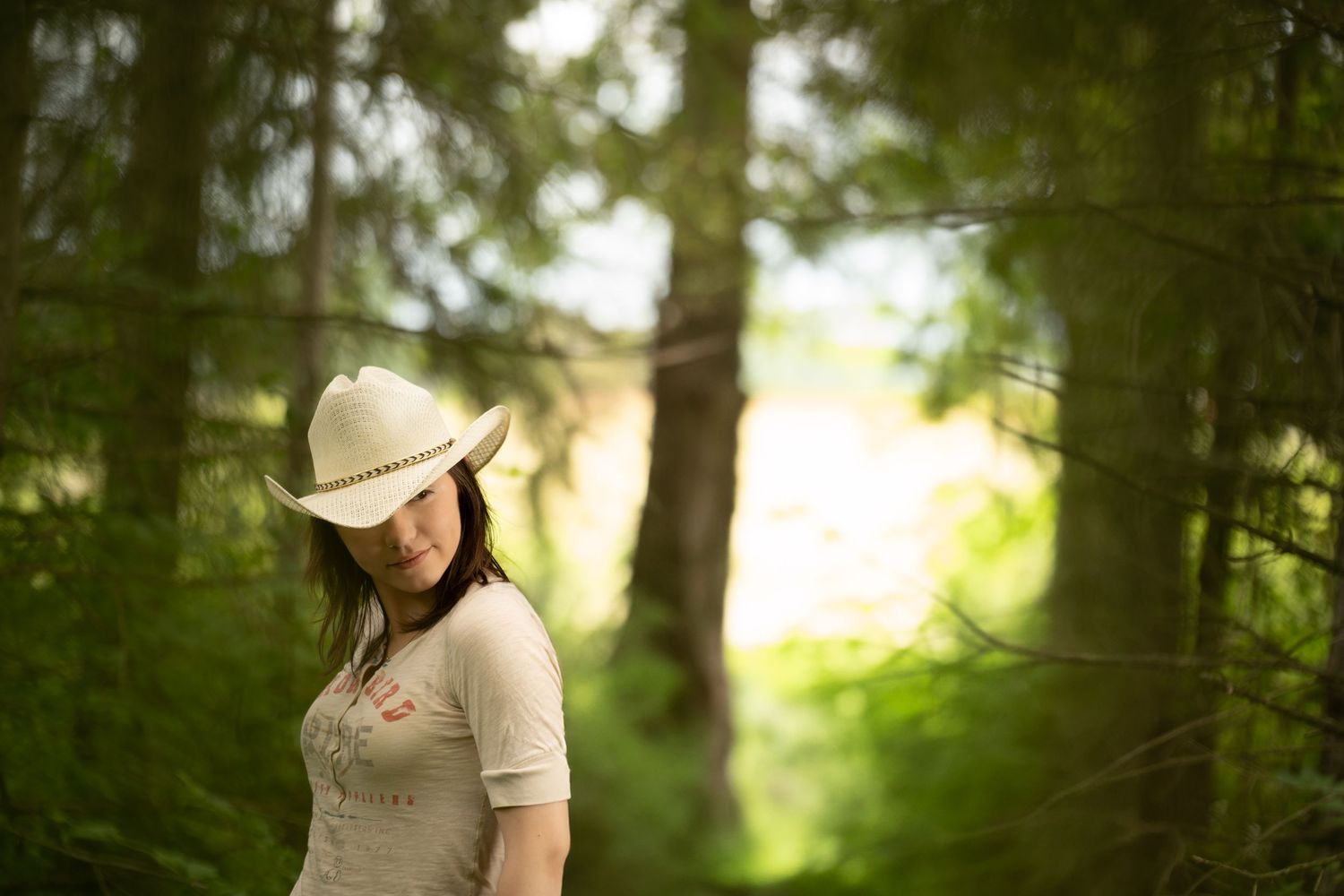A young woman in a white cowboy hat and beige top stands thoughtfully in a lush green forest setting.