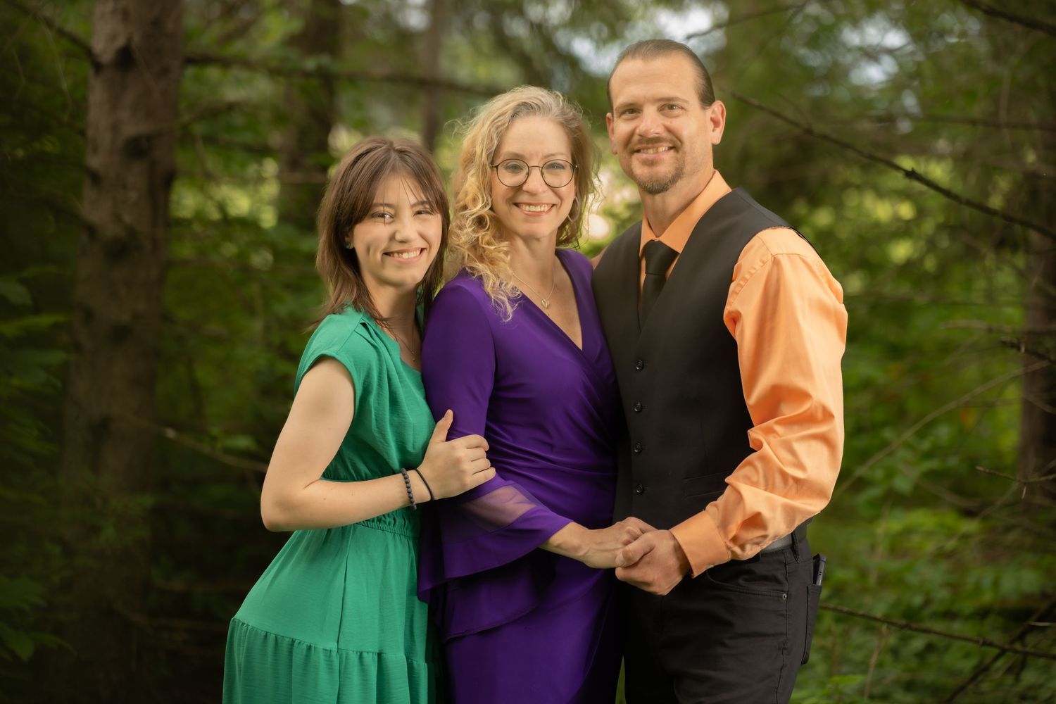 Three smiling people posing outdoors in a forest setting, wearing colorful formal attire for a family portrait.