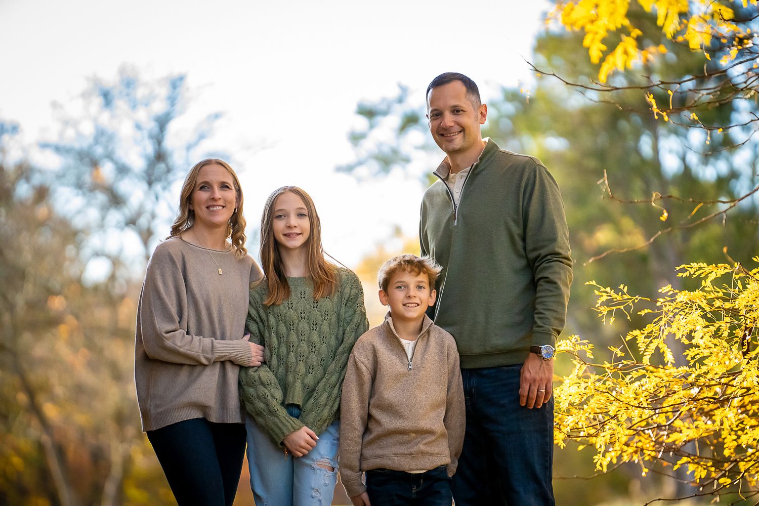 A family of four poses together outdoors surrounded by vibrant yellow autumn foliage.