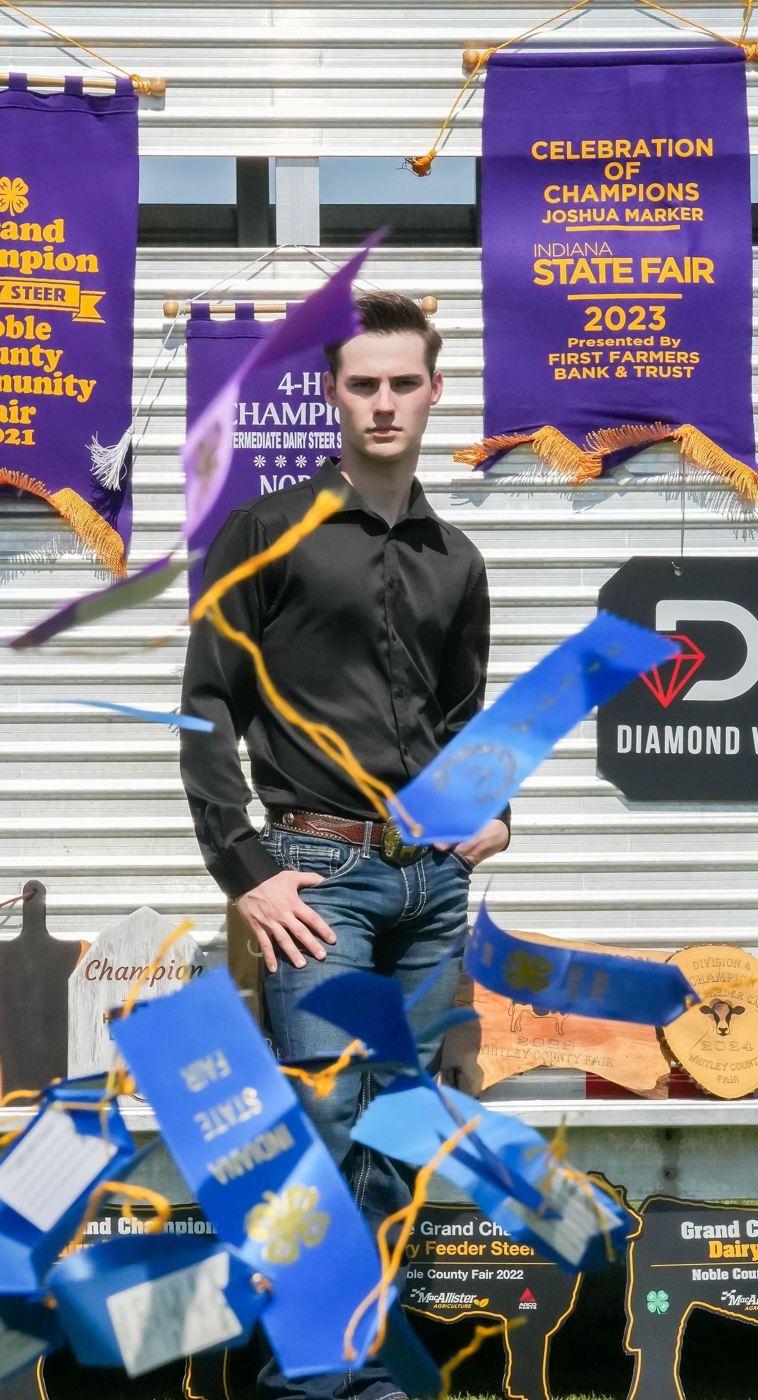 Young man in black shirt standing before 4-H state fair champion banners and blue ribbons.