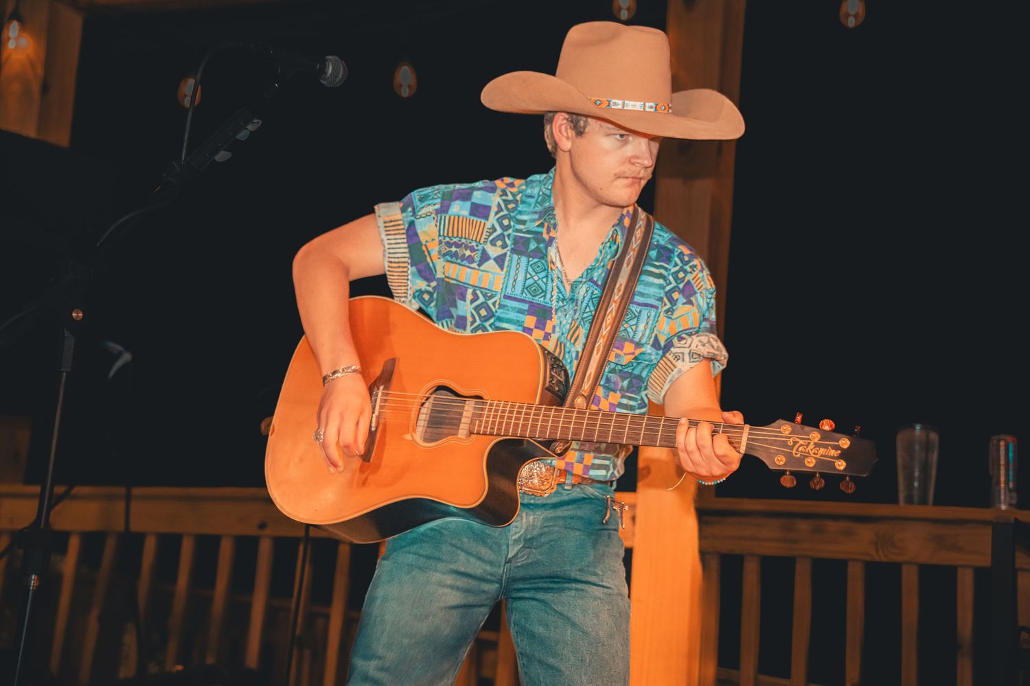Country guitarist in tan cowboy hat and colorful Hawaiian shirt plays acoustic guitar on warmly lit stage.