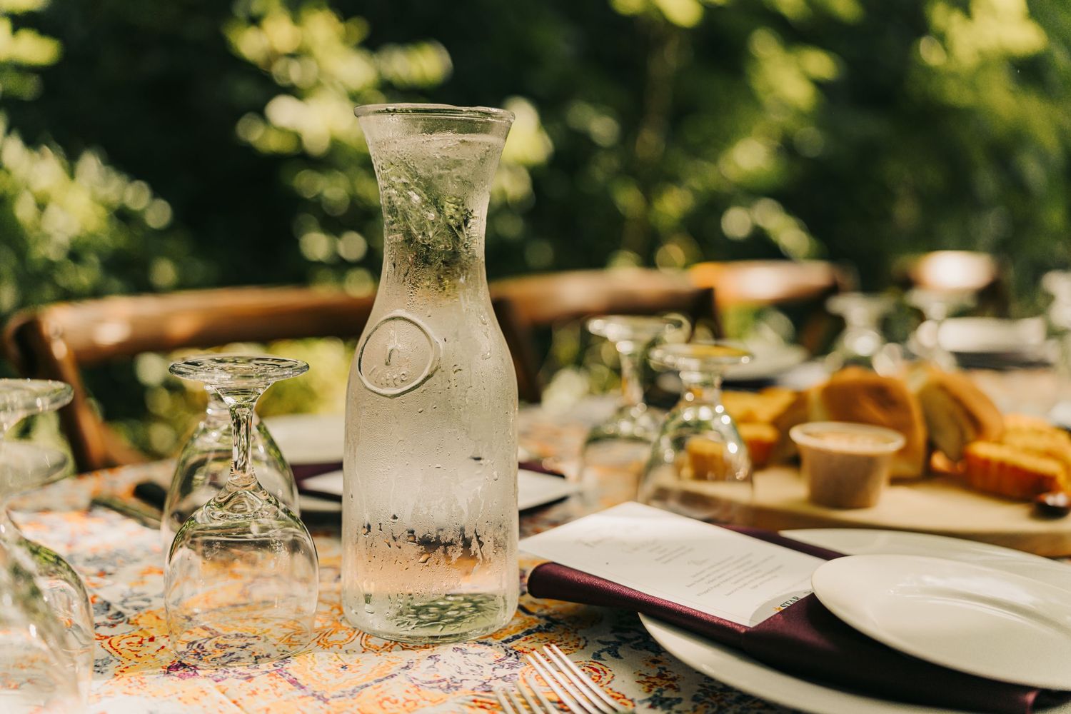 Glass carafe of water with lemon on a rustic outdoor dining table surrounded by greenery.