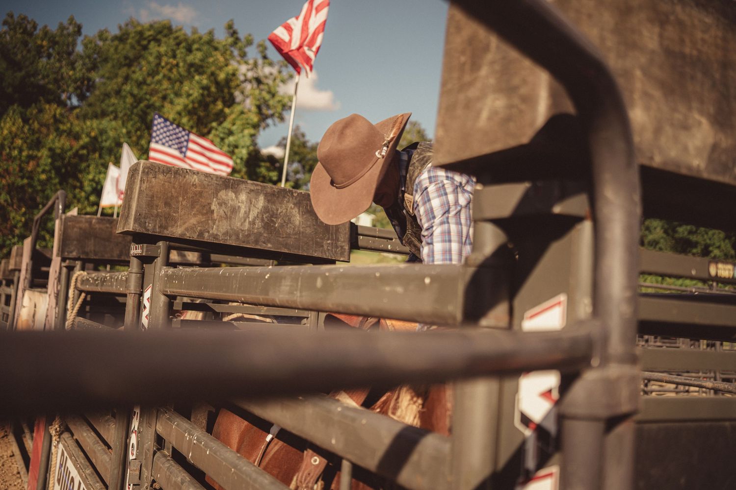 American flags wave above rodeo chutes with a cowboy hat visible behind metal gate fencing.