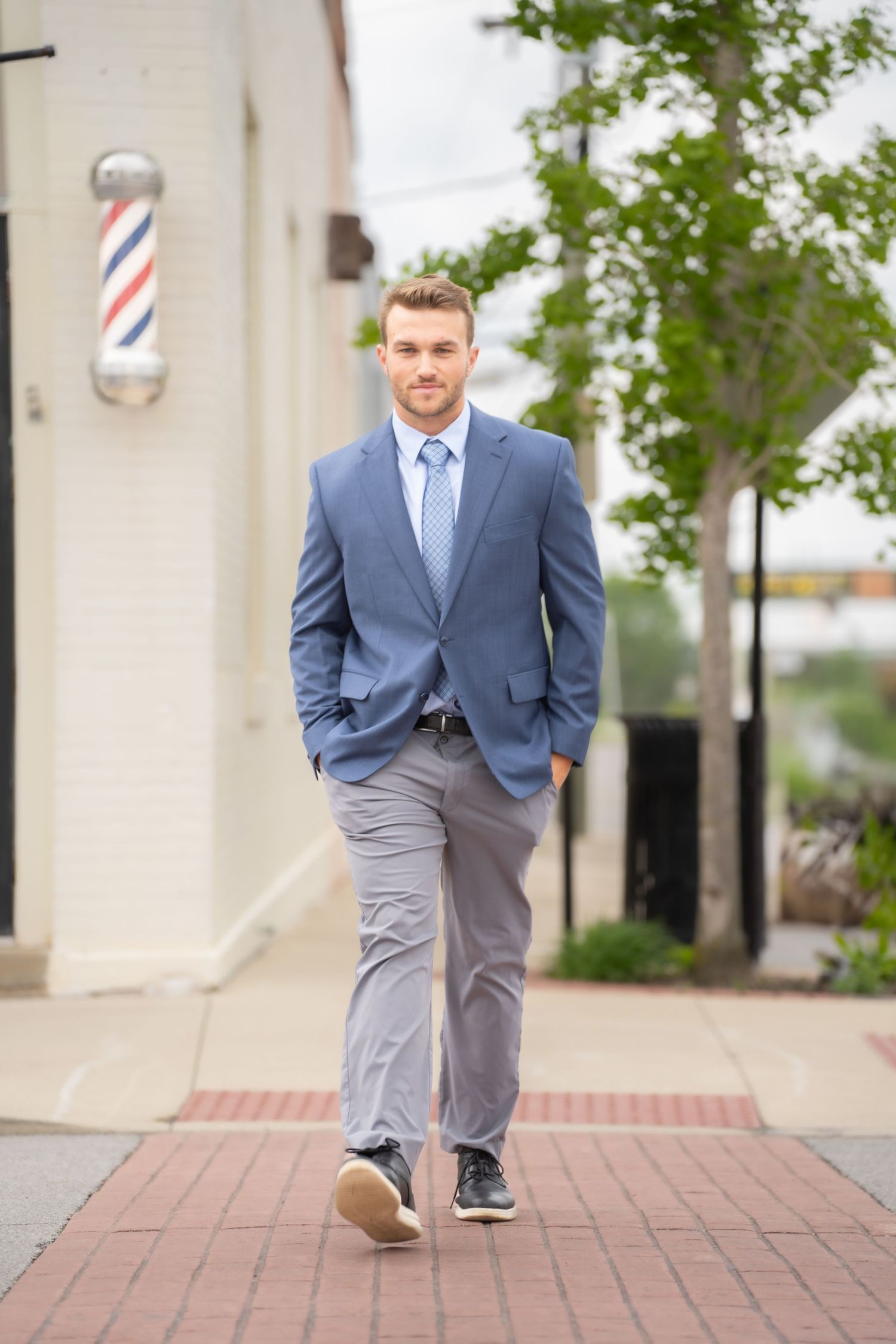 A confident businessman in a blue suit and tie walks along a brick sidewalk near a barbershop.