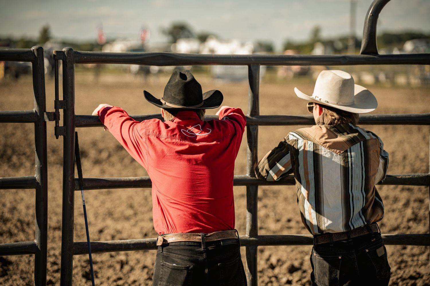 Two cowboys in hats lean on a metal gate overlooking a dirt rodeo arena.