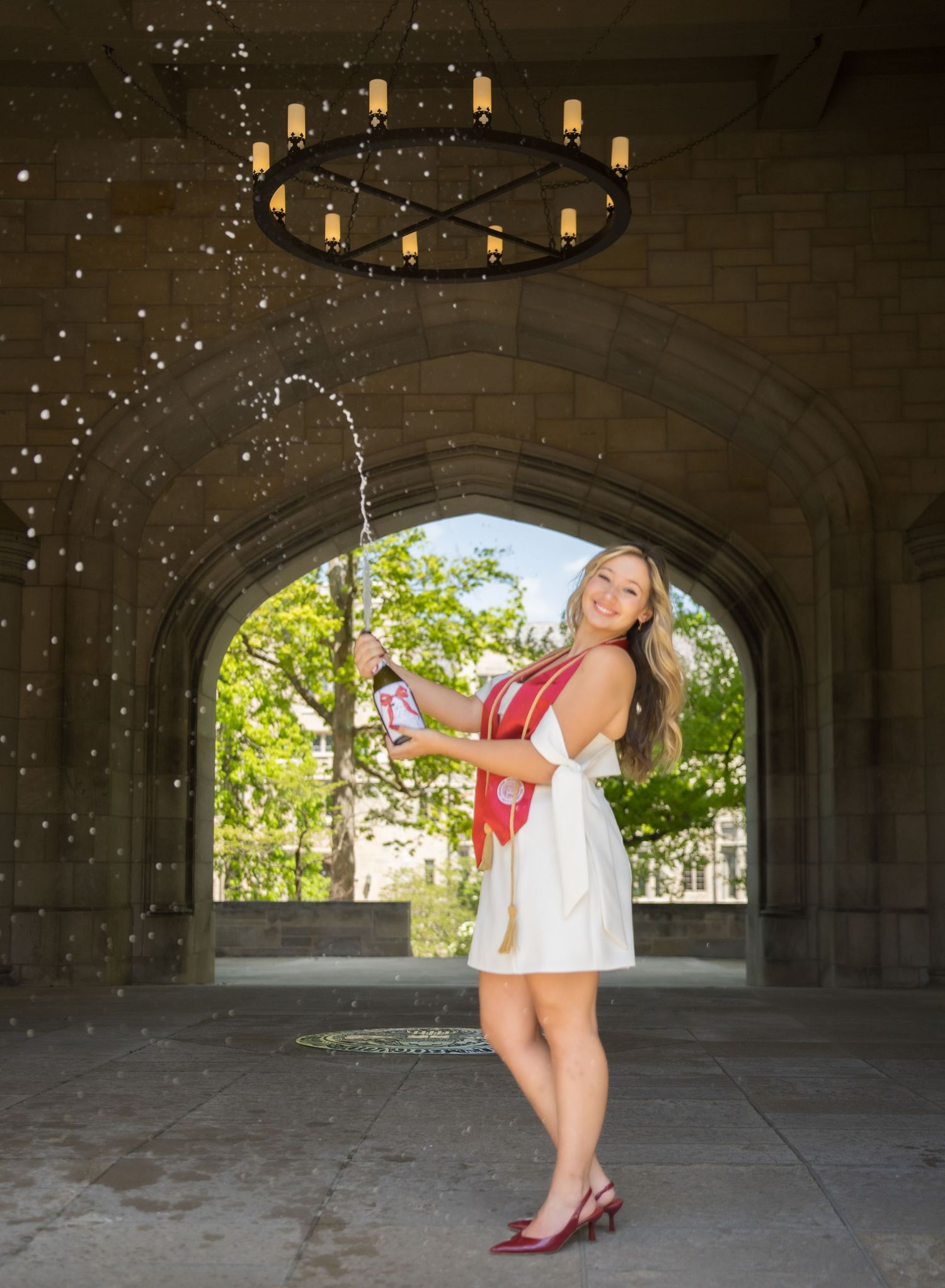 Woman in white dress popping champagne bottle under stone archway with chandelier overhead.