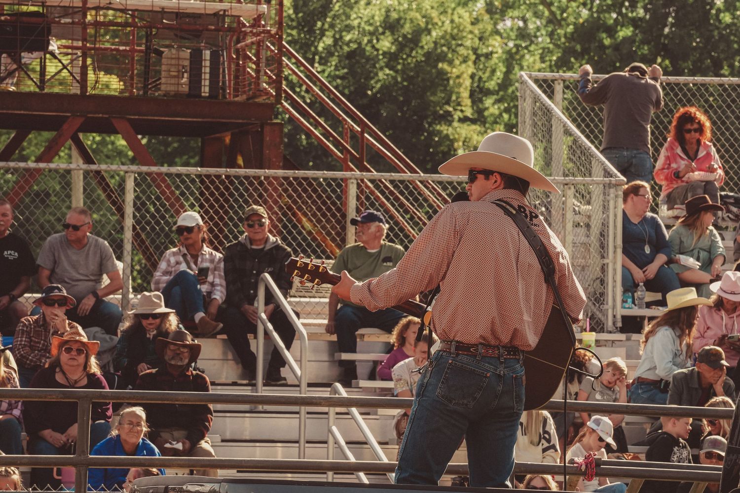 Cowboy performer engaging with crowd at outdoor rodeo grandstand on sunny afternoon.