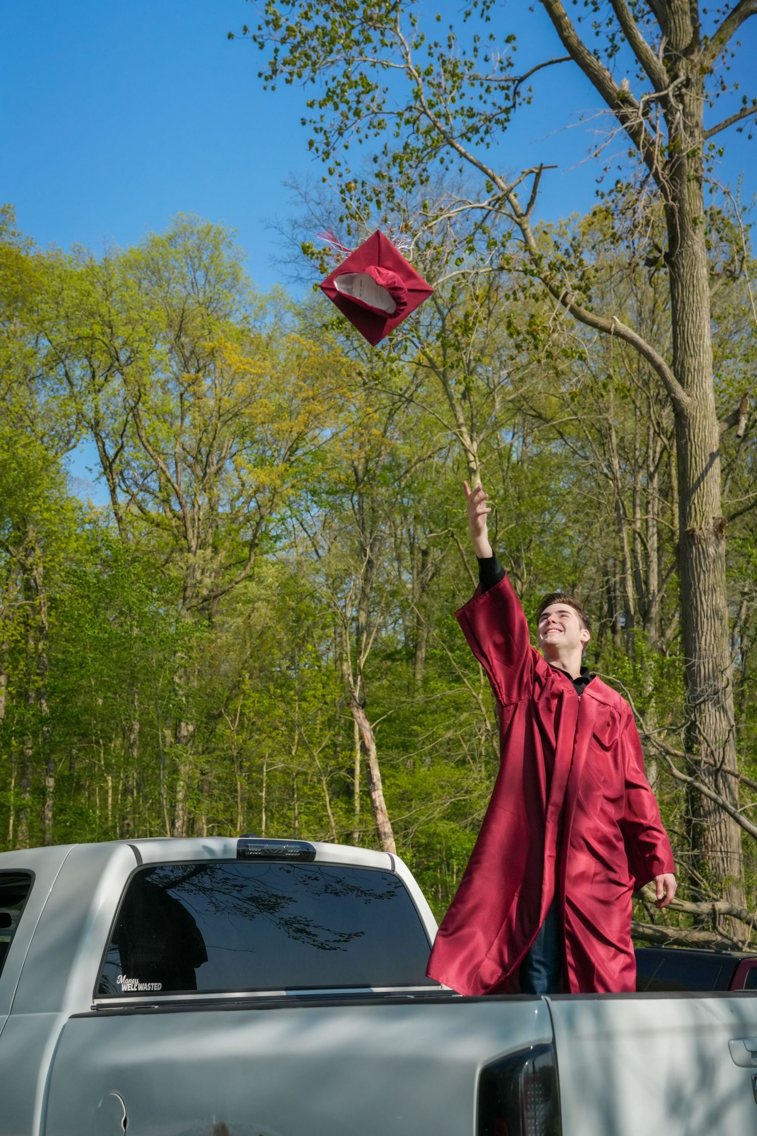 Smiling graduate in red cap and gown tosses graduation cap from truck bed surrounded by trees.