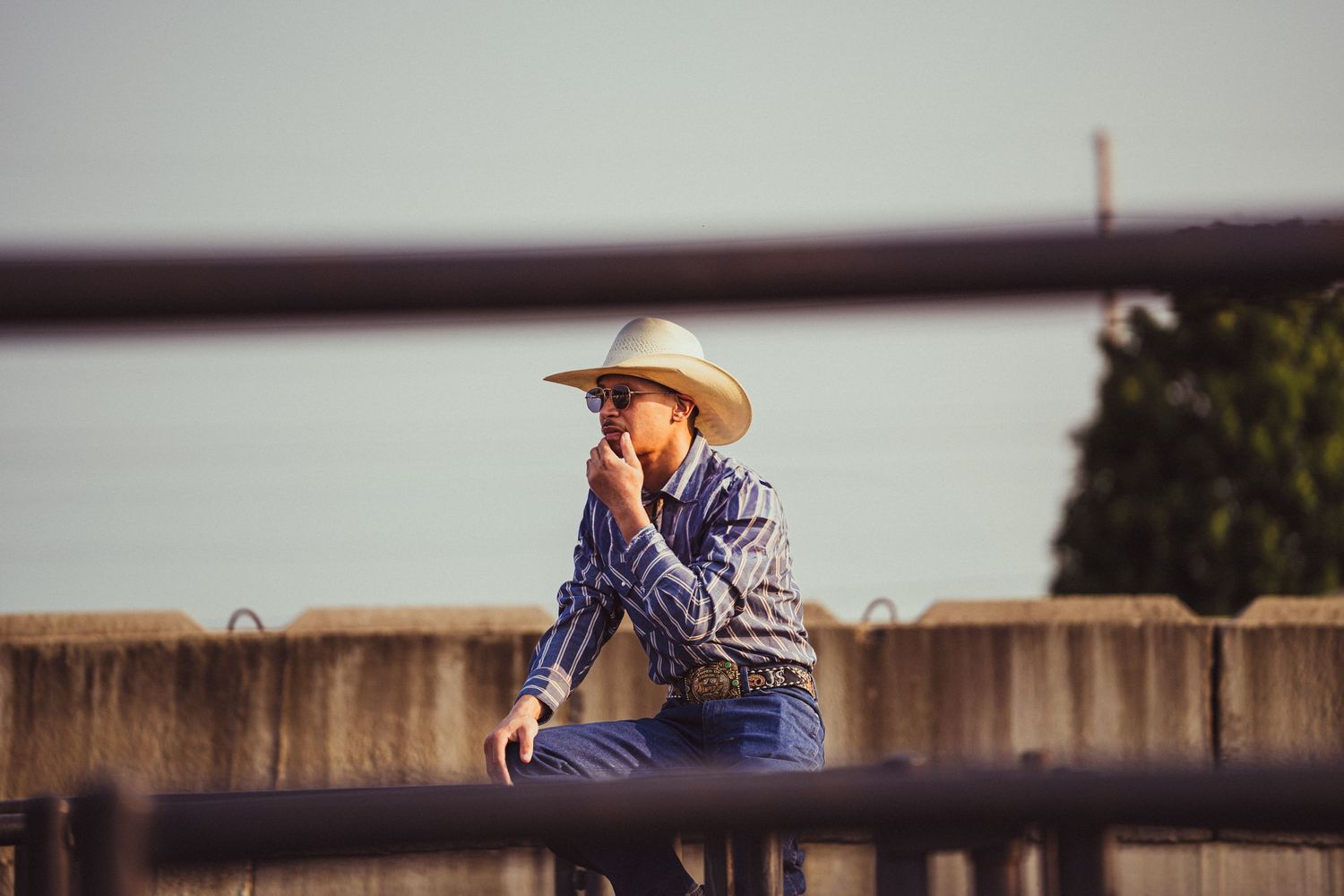 A cowboy in a striped shirt and hat sits on a fence, thoughtfully touching his face at an outdoor rodeo.