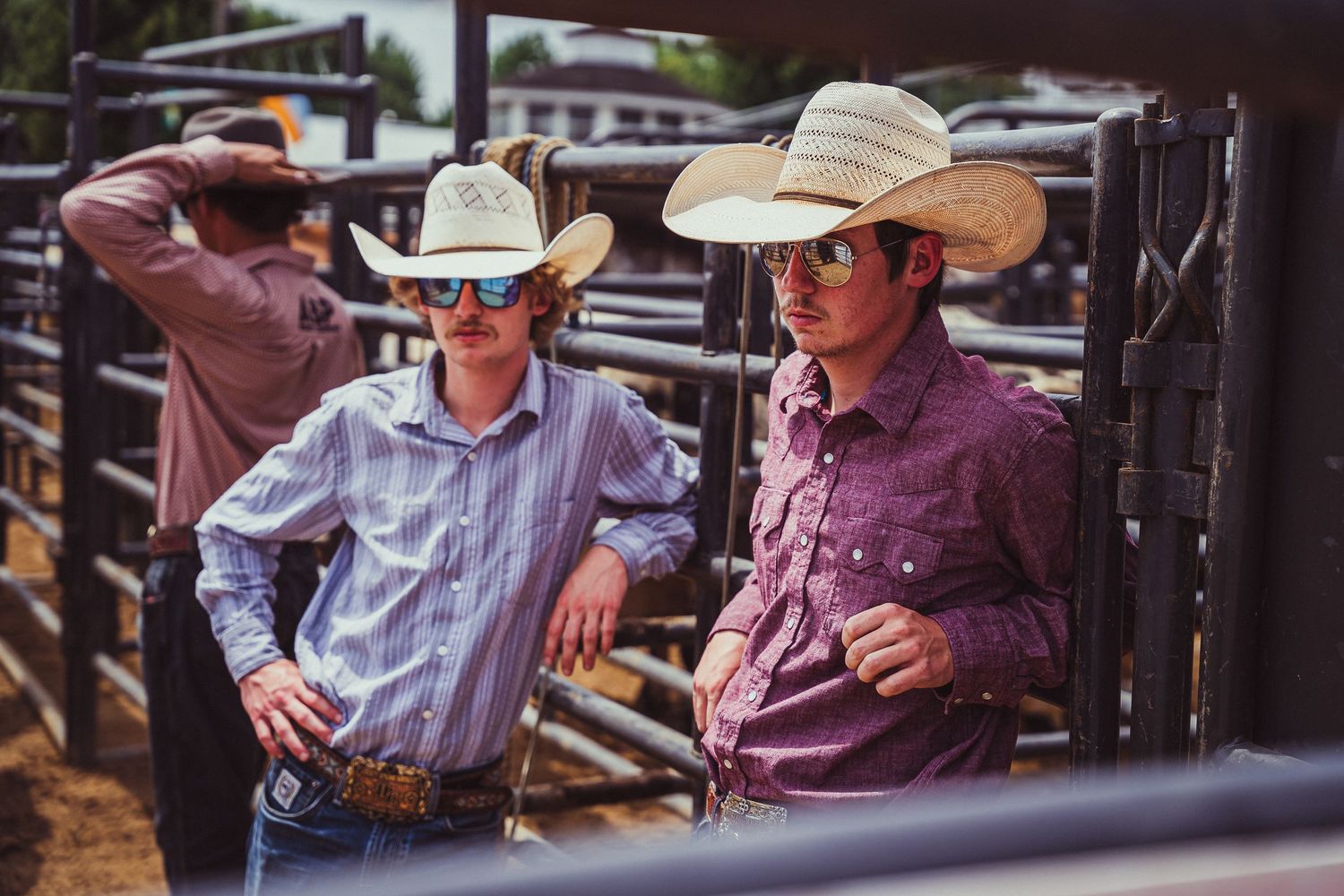 Two cowboys in white hats and sunglasses stand by metal rodeo pens in western shirts.
