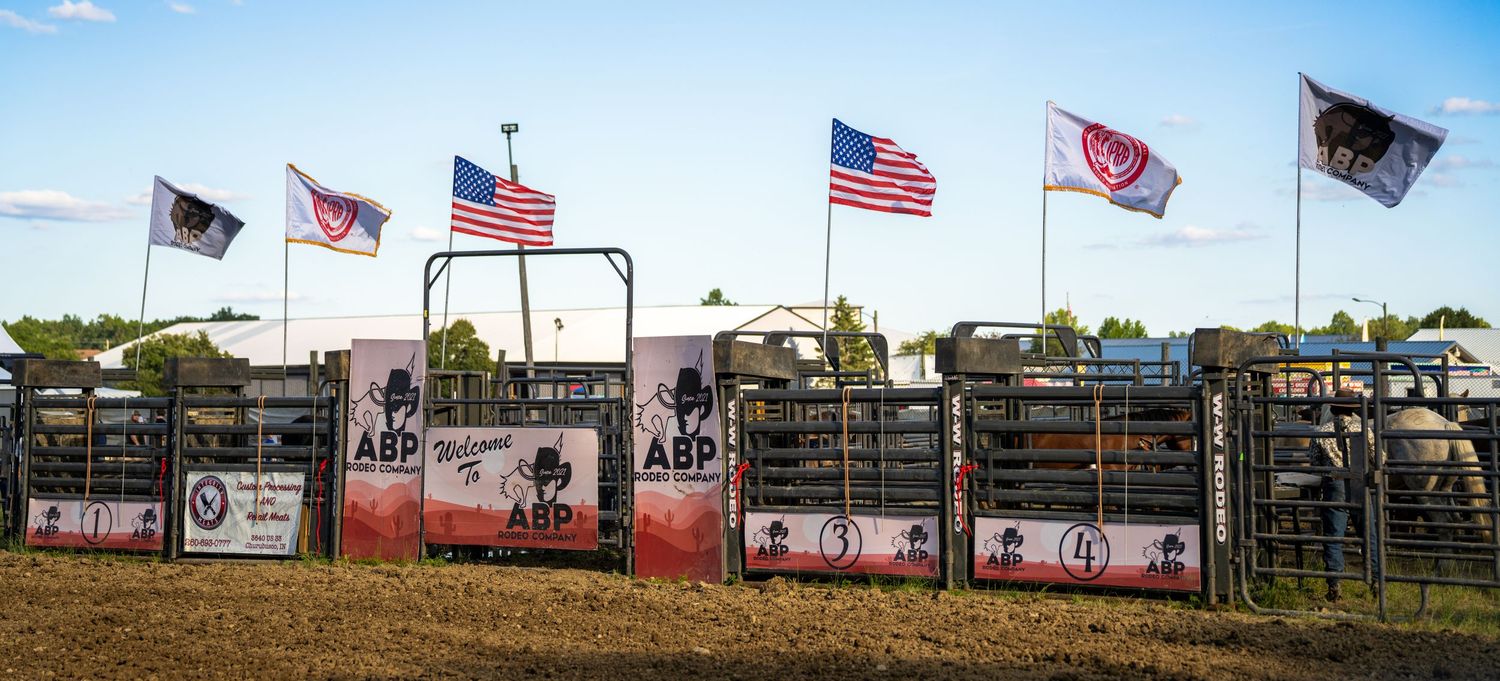 ABP rodeo arena with American and state flags flying above metal chutes and sponsor banners at an outdoor bull riding event.